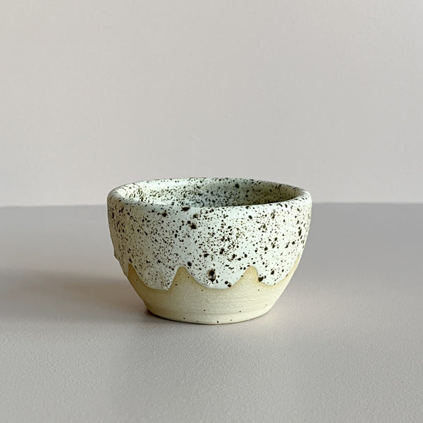 A mini ceramic bowl in a natural clay color featuring an off-white and brown speckled glaze with a wavy edge, sat against a neutral backdrop.