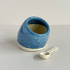 A ceramic salt cellar featuring a rich cornflower blue glaze and a natural clay base, with a small wood spoon resting in front, all against a neutral backdrop.
