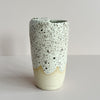 A ceramic travel cup featuring an off-white and brown speckled glaze with a wavy finish near the bottom, revealing the natural clay beneath. The cup sits against a neutral backdrop.