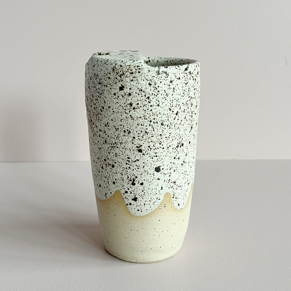 A ceramic travel cup featuring an off-white and brown speckled glaze with a wavy finish near the bottom, revealing the natural clay beneath. The cup sits against a neutral backdrop.