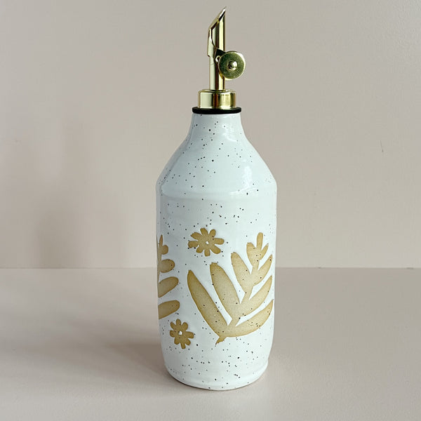 A large ceramic oil pourer with a gold spout. The cylindrical cruet sports a bright white speckled glaze with a floral resist design, allowing the natural clay to show through. The bottle sits against a neutral backdrop.