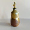 A ceramic oil cruet bottle in an earthy brown and red shino glaze with a gold pour spout against a neutral backdrop.