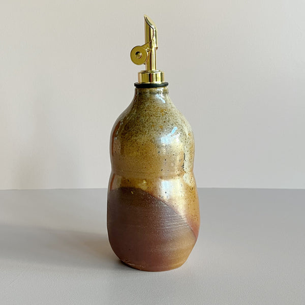 A ceramic oil cruet bottle in an earthy brown and red shino glaze with a gold pour spout against a neutral backdrop.