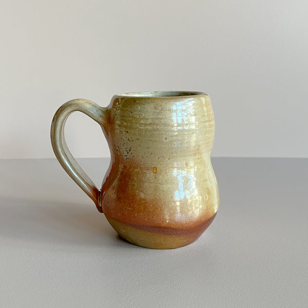 A bubble shaped ceramic mug with an earthy tan and red-brown shino glaze against a neutral backdrop.