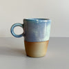 A ceramic mug with a loop shaped handle. It's dipped in a wood-fired speckly blue-purple glaze with the bottom half of the mug revealing the natural clay color. It sits against a neutral backdrop.