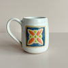 A white speckled glazed ceramic mug with a floral quilt square tile design in blue, red-orange, and bright green, set against a neutral backdrop.