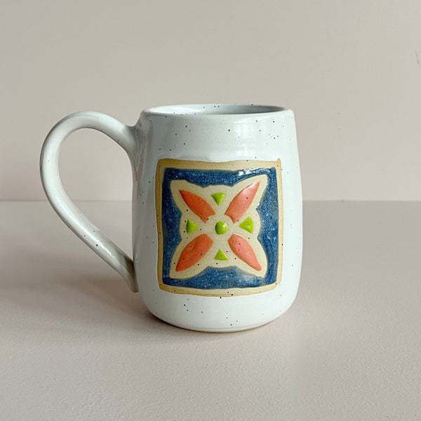 A white speckled glazed ceramic mug with a floral quilt square tile design in blue, red-orange, and bright green, set against a neutral backdrop.
