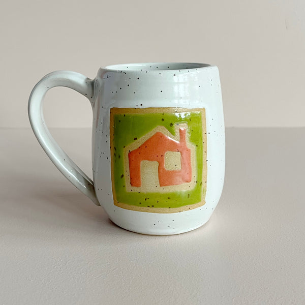 A ceramic mug in a white speckled glaze featuring a tile design of a red-orange house silhouette surrounded by bright green and natural clay colors. The mug sits against a neutral backdrop.