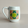 A speckled white glazed ceramic mug with a pink, green, and teal quilt square glaze design set against a neutral backdrop.