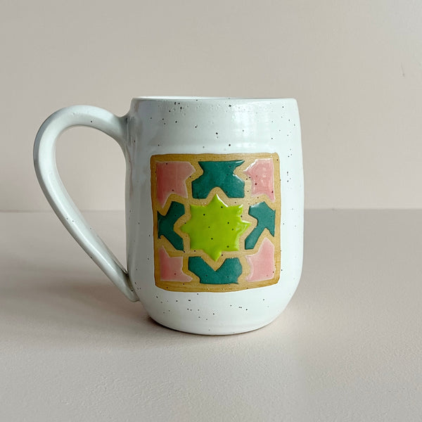 A speckled white glazed ceramic mug with a pink, green, and teal quilt square glaze design set against a neutral backdrop.