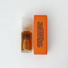 A small clear bottle of fragrance oil with a translucent cap stands next to a bright orange rectangular box labeled with ingredients on a clean white background. The bottle label reads 'STRANGE DREAMER'.