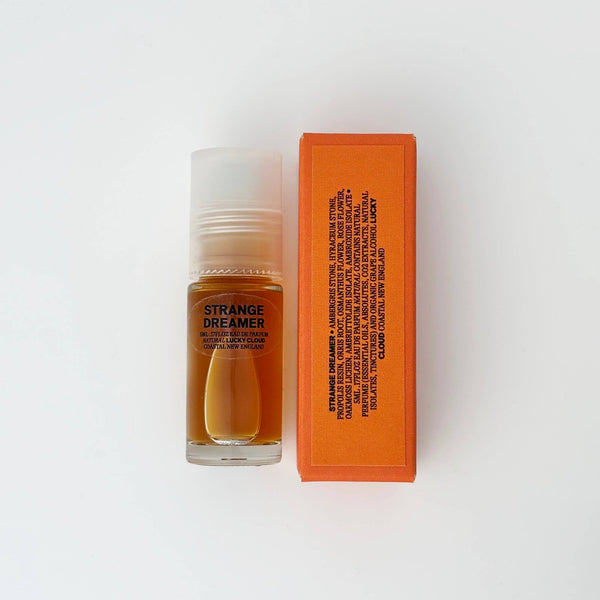 A small clear bottle of fragrance oil with a translucent cap stands next to a bright orange rectangular box labeled with ingredients on a clean white background. The bottle label reads 'STRANGE DREAMER'.