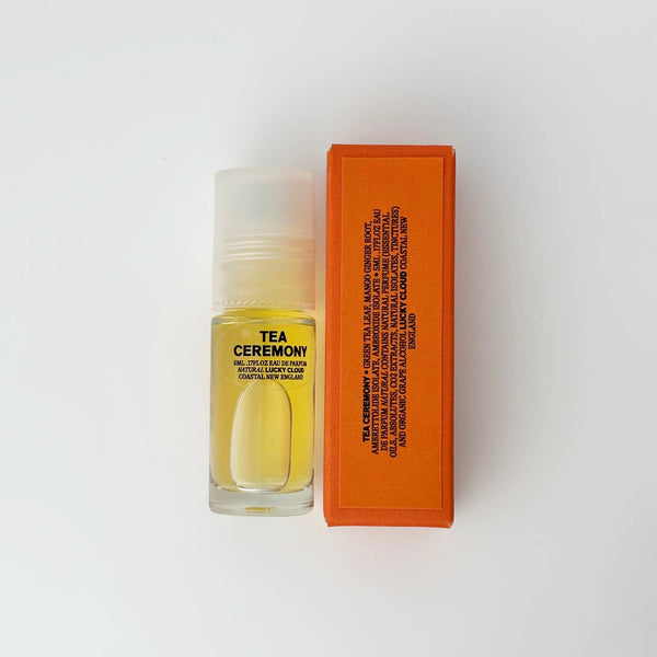 A small clear roll-on bottle filled with pale yellow natural labeled 'TEA CEREMONY' stands next to a bright orange rectangular box labeled with ingredients on a clean white background.