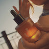 A close-up shot of a person holding a frosted amber glass dropper bottle with a black dropper cap. The bottle features a stylized white logo and is illuminated by warm golden light, with a blurred outdoor background.