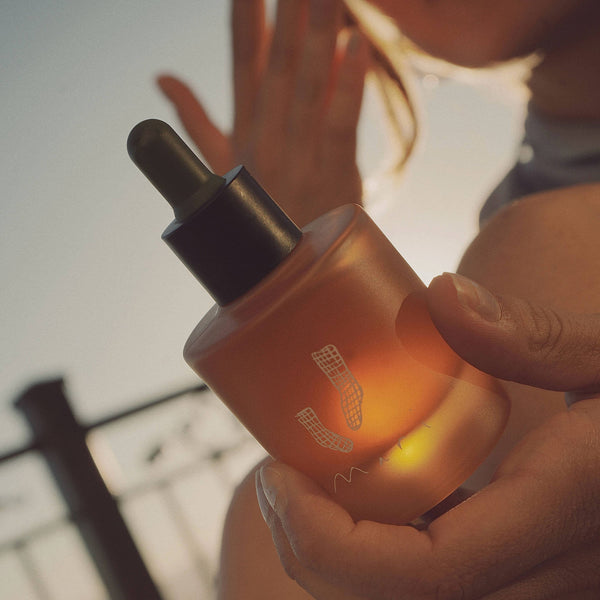 A close-up shot of a person holding a frosted amber glass dropper bottle with a black dropper cap. The bottle features a stylized white logo and is illuminated by warm golden light, with a blurred outdoor background.