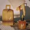 A hand in a cream knit sleeve reaches toward a small amber dropper bottle labeled MARI, next to a matching kraft paper gift box with the same logo, on a textured surface with warm directional sunlight and shadows.