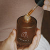 Hand holding a brown glass bottle with a dropper labeled 'mari' against a neutral background