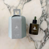 A dark glass oil bottle with dropper and light blue paper packaging on a marble surface.
