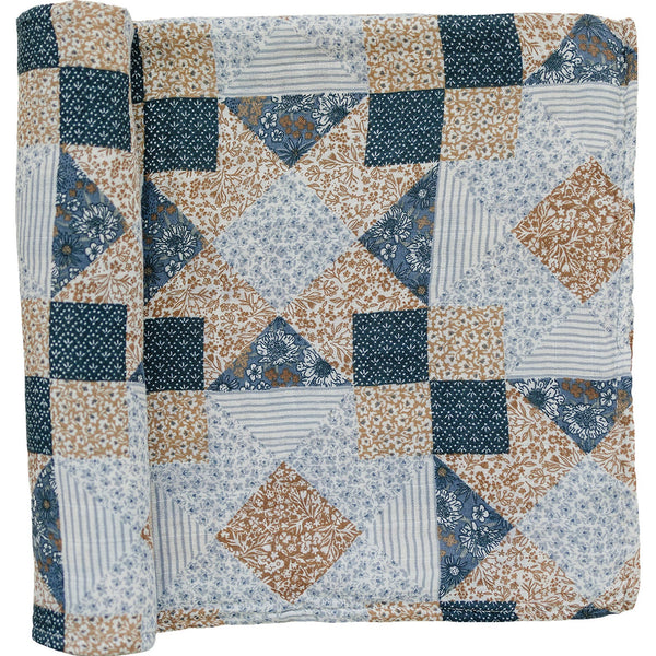 A partially unrolled muslin swaddle blanket with a blue, brown and white patchwork quilt print on a white backdrop.