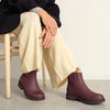 A person sitting in a chair wearing light yellow pants and a dark jacket with dark red pull-on ankle boots against a concrete floor and white wall.