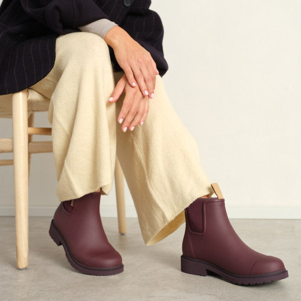 A person sitting in a chair wearing light yellow pants and a dark jacket with dark red pull-on ankle boots against a concrete floor and white wall.
