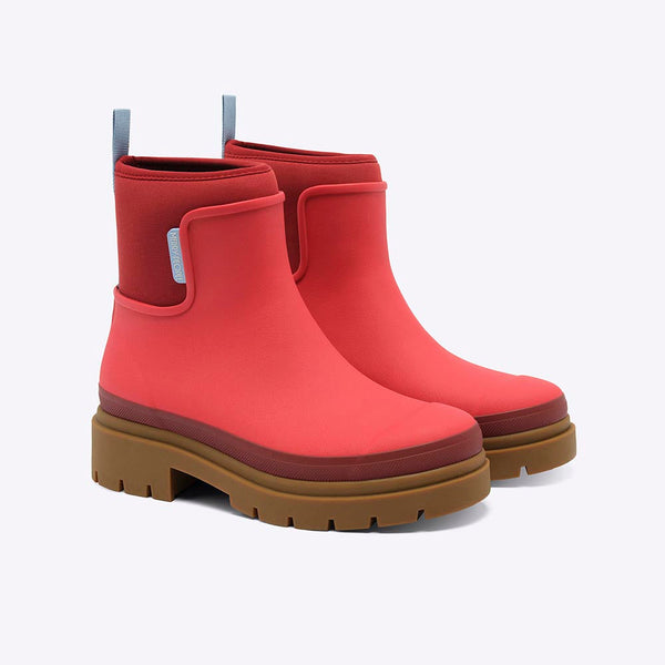 A pair of bright chilli red neoprene rainboots with light blue tab accents and a light brown chunky low heeled sole against a white backdrop.