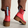 A person wearing brown raw hem pants with a pair of bright chilli red and light blue accent rainboots with a chunky light brown sole against a neutral backdrop.