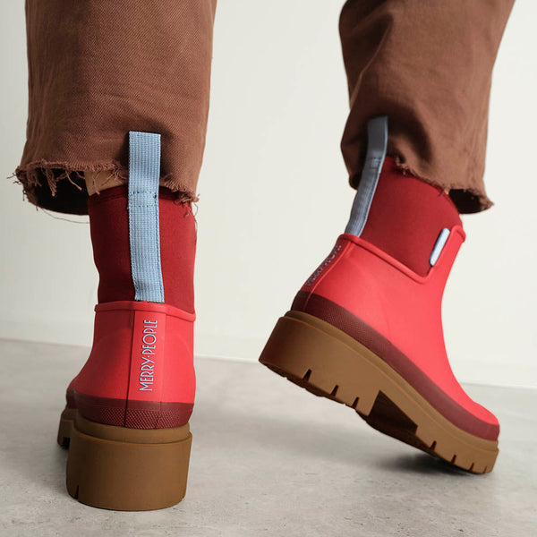 A person wearing brown raw hem pants with a pair of bright chilli red and light blue accent rainboots with a chunky light brown sole against a neutral backdrop.