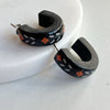 A pair of glossy black polymer clay earrings in a chunky half hoop shape, hand-painted with Nordic-inspired floral designs in orange and white, resting against a marble surface.