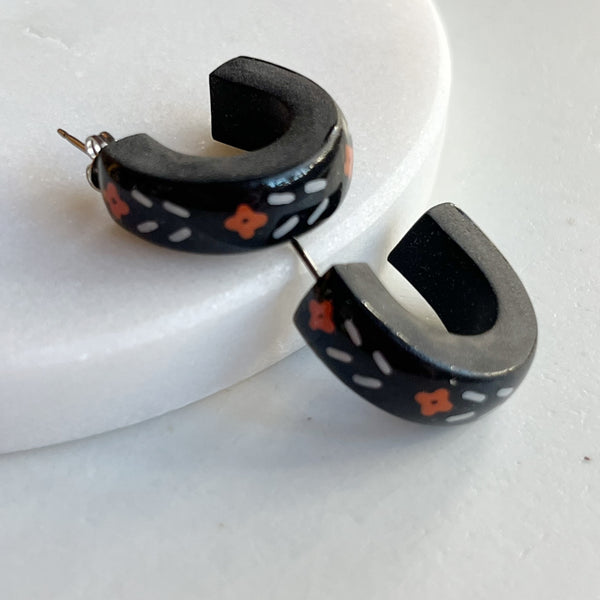 A pair of glossy black polymer clay earrings in a chunky half hoop shape, hand-painted with Nordic-inspired floral designs in orange and white, resting against a marble surface.