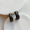 A pair of glossy black polymer clay earrings in a chunky half hoop shape, hand-painted with Nordic-inspired floral designs in orange and white, resting against a marble surface.