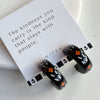 A pair of glossy black polymer clay earrings in a chunky half hoop shape, hand-painted with Nordic-inspired floral designs in orange and white on their white brand card, resting against a marble surface.
