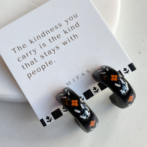 A pair of glossy black polymer clay earrings in a chunky half hoop shape, hand-painted with Nordic-inspired floral designs in orange and white on their white brand card, resting against a marble surface.
