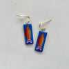 A pair of glossy blue polymer clay earrings in a long rectangular shape hand=painted with a colorful fish, hanging from gold hooks on a neutral background.