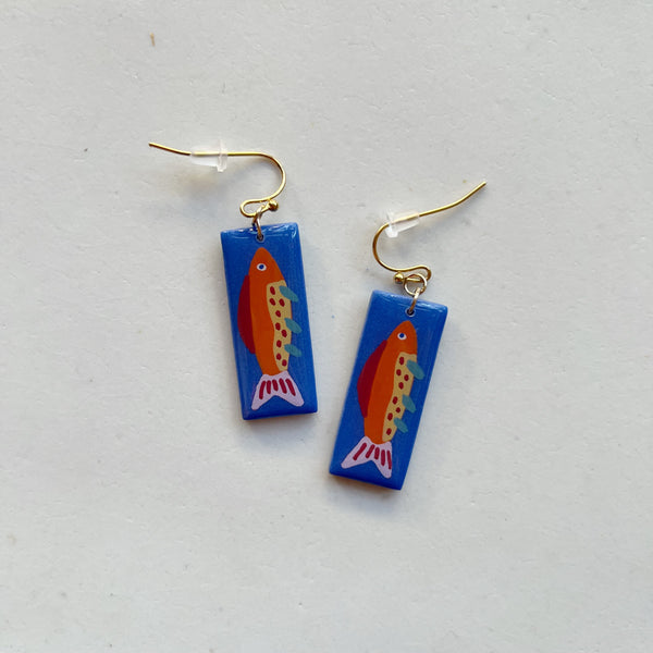 A pair of glossy blue polymer clay earrings in a long rectangular shape hand=painted with a colorful fish, hanging from gold hooks on a neutral background.