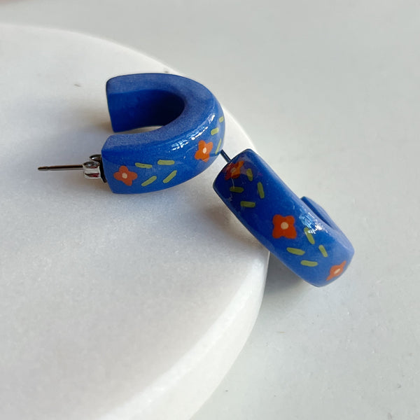 A pair of glossy blue polymer clay earrings in a chunky half hoop shape hand-painted with a Nordic-inspired floral design in red and green, resting against a marble surface.