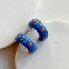 A pair of glossy blue polymer clay earrings in a chunky half hoop shape hand-painted with a Nordic-inspired floral design in red and green, resting against a marble surface.