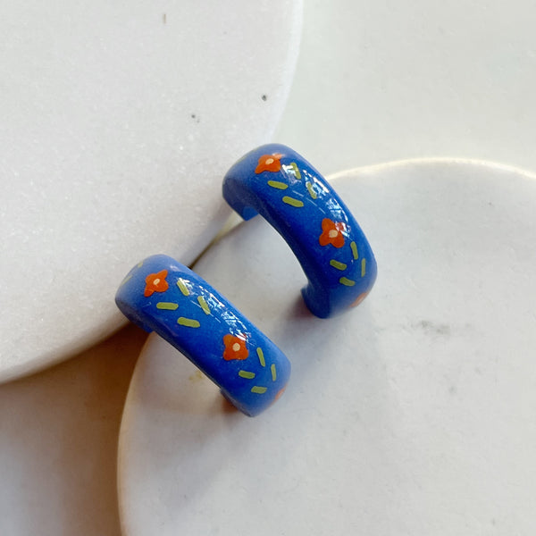 A pair of glossy blue polymer clay earrings in a chunky half hoop shape hand-painted with a Nordic-inspired floral design in red and green, resting against a marble surface.