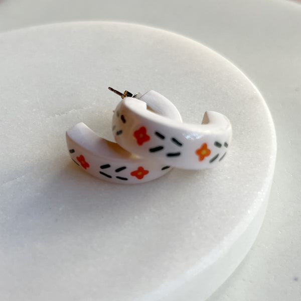 A pair of glossy ivory polymer clay earrings in a chunky half hoop, hand-painted with Nordic-inspired floral motifs in orange and dark green, stacked on top of each other resting against a marble surface.