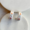 A pair of glossy ivory polymer clay earrings in a chunky half hoop, hand-painted with Nordic-inspired floral motifs in orange and dark green, resting against a marble backdrop.