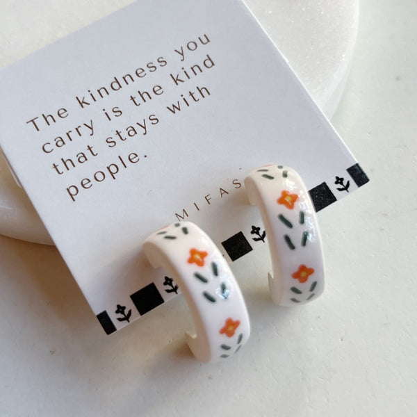 A pair of glossy ivory polymer clay earrings in a chunky half hoop, hand-painted with Nordic-inspired floral motifs in orange and dark green, on their white branded card, resting against a marble backdrop.