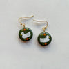 A pair of glossy olive green polymer clay earrings in a circle shape hand-painted with a sweet duck and flower design, hanging from gold hooks. 