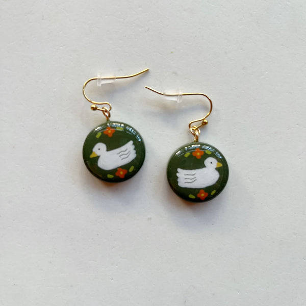 A pair of glossy olive green polymer clay earrings in a circle shape hand-painted with a sweet duck and flower design, hanging from gold hooks. 