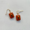 A pair of glossy terracotta polymer clay earrings in mini rectangles hand-painted with a dainty yellow and green flower, hanging from gold hooks on a neutral backdrop.