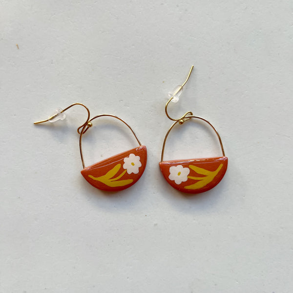 A pair of glossy terracotta polymer clay earrings in a half circle shape hand-painted with a white and chartreuse floral motif, hanging from gold wire arcs and gold hooks. 