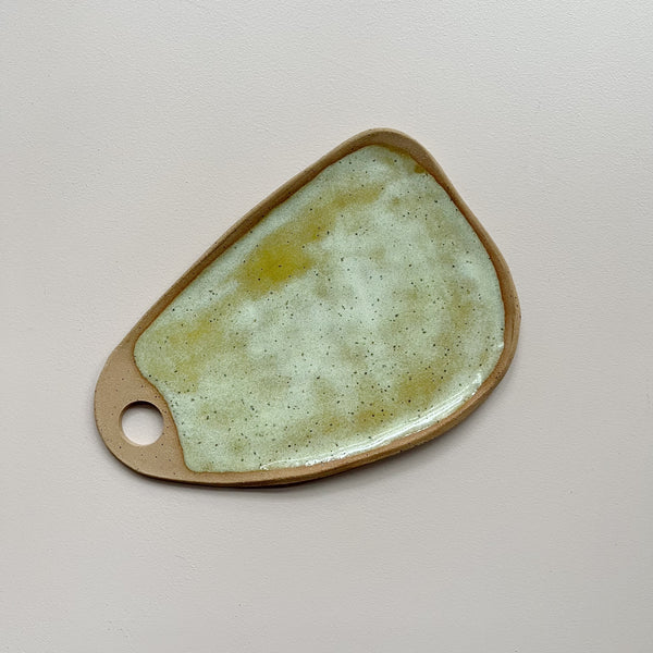 A ceramic serving board with a circular cutout handle and swirling golden yellow and off-white speckly glaze in an oblong wedge shape against a neutral backdrop.