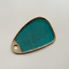 A ceramic serving board with a circular cutout handle and a vibrant teal speckled glaze in an oblong wedge shape against a neutral backdrop.