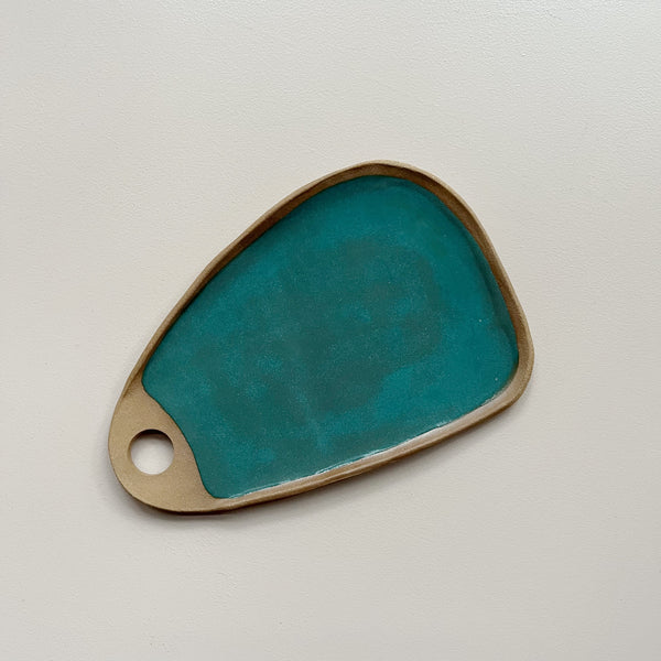A ceramic serving board with a circular cutout handle and a vibrant teal speckled glaze in an oblong wedge shape against a neutral backdrop.