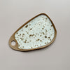 A ceramic serving board with a circular cutout handle and bright white glaze with brown speckles in an oblong wedge shape against a neutral backdrop.