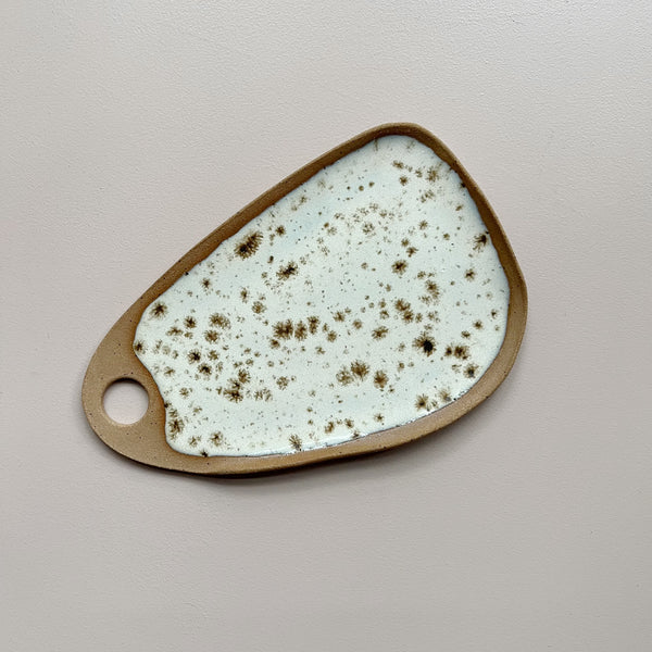 A ceramic serving board with a circular cutout handle and bright white glaze with brown speckles in an oblong wedge shape against a neutral backdrop.